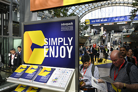 Un stand ferial de Interpack Alliance con un gran letrero azul y amarillo en el que se lee «SIMPLY ENJOY». En un expositor se muestran folletos con el logotipo, y al fondo se ven visitantes en el evento.