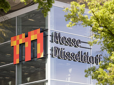El exterior de un moderno edificio acristalado con el logotipo «Messe Düsseldorf» destacado en la fachada, parcialmente enmarcado por las ramas verdes de los árboles.